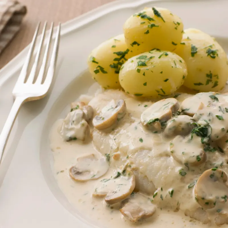 Fricasseed white fish fillet topped with creamy mushroom herb sauce, served with parsley potatoes on a white plate.