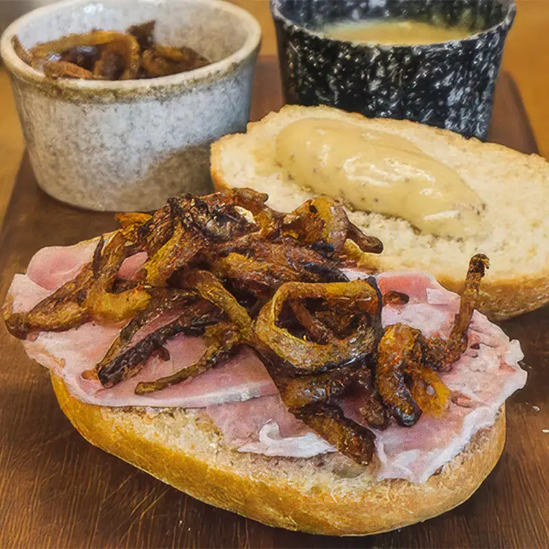 Ham sandwich topped with crispy fried onions and mustard on a wooden board, with condiments in the background.