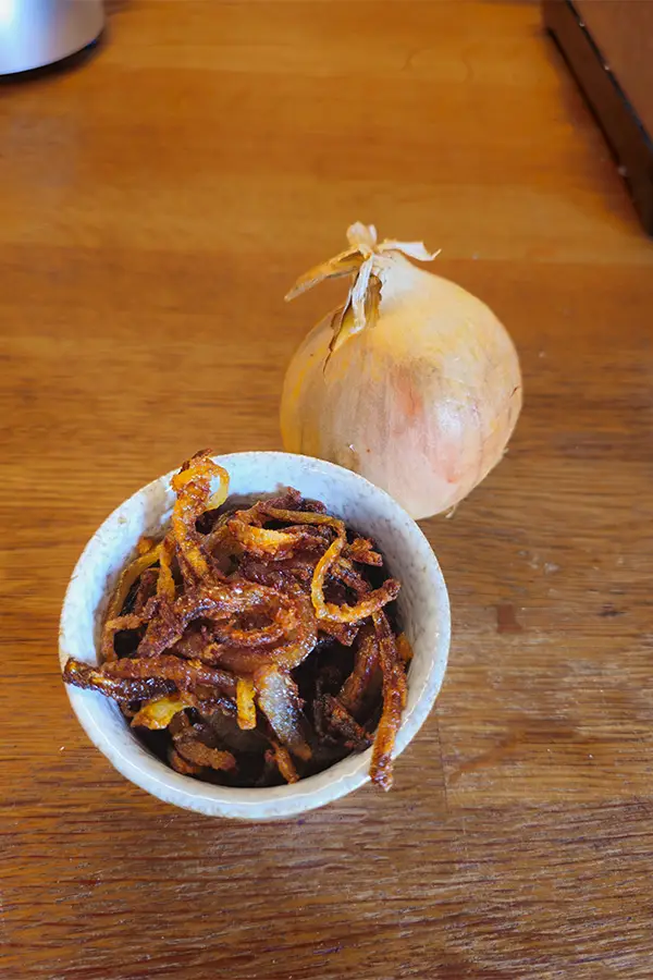 Crispy homemade German fried onions served in a small ceramic bowl next to a whole yellow onion on a wooden surface.