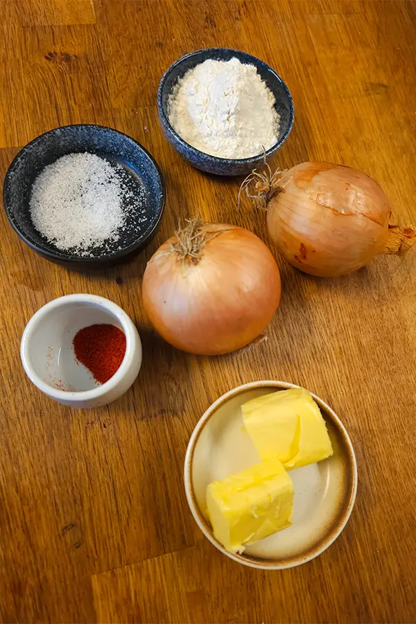 Overhead view of fried onion ingredients: yellow onions, flour, salt, paprika, and butter arranged on a wooden surface.