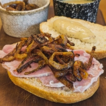 Ham sandwich topped with crispy fried onions and mustard on a wooden board, with condiments in the background.