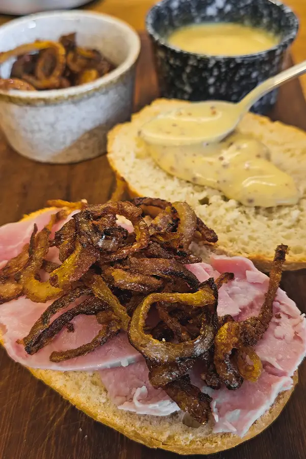 Close-up of a ham sandwich being assembled with fried onions and mustard spooned onto the top bun.