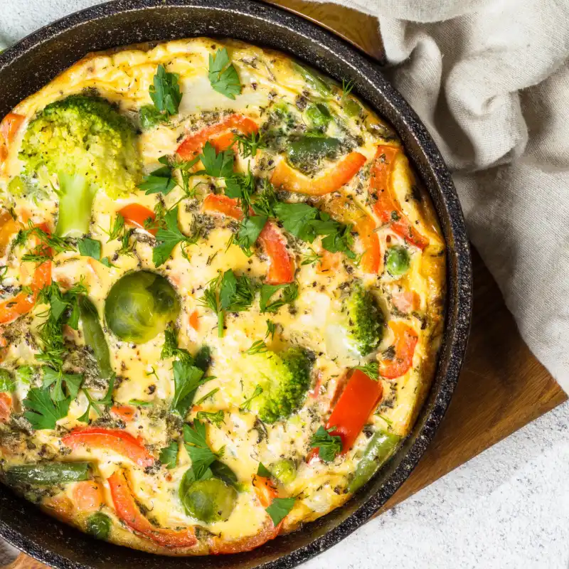 Colorful vegetable frittata in a black skillet, filled with broccoli, Brussels sprouts, red peppers, and herbs.