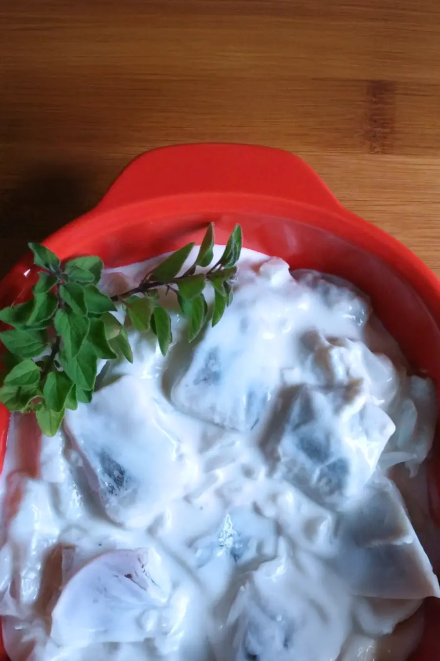 A red bowl containing herring salad with a fresh sprig of green leaves on top, placed on a wooden surface.