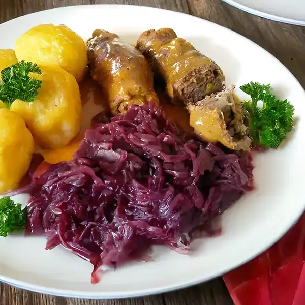 Traditional Rinderrouladen with gravy, red cabbage, and potato dumplings on a white plate.