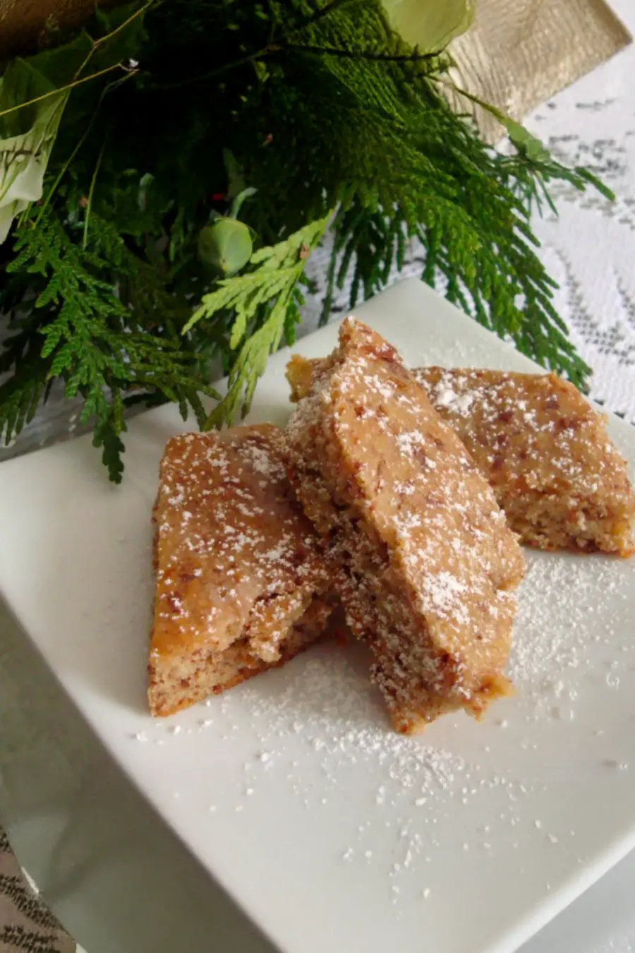 Three powdered sugar-coated dessert bars stacked on a square white plate, with greenery in the background.