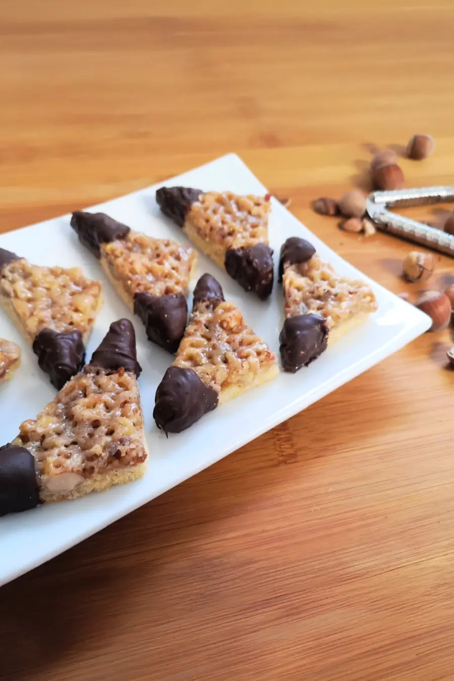 A plate of triangular nut bars partially dipped in chocolate, placed on a wooden surface with some scattered nuts and a nutcracker beside it.