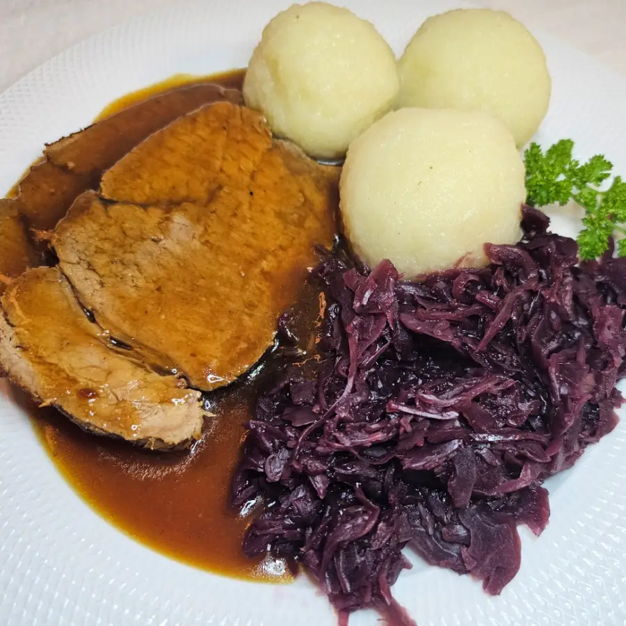 Sauerbraten with gravy, red cabbage, and German potato dumplings on a dinner plate.