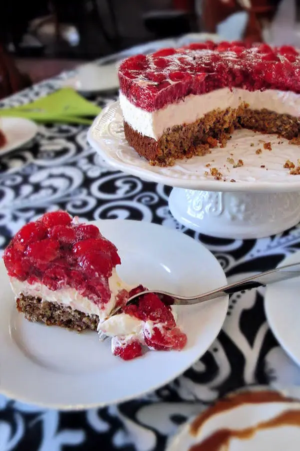 Slice of raspberry torte on a plate next to a cake stand with the remaining cheesecake on a patterned tablecloth.
