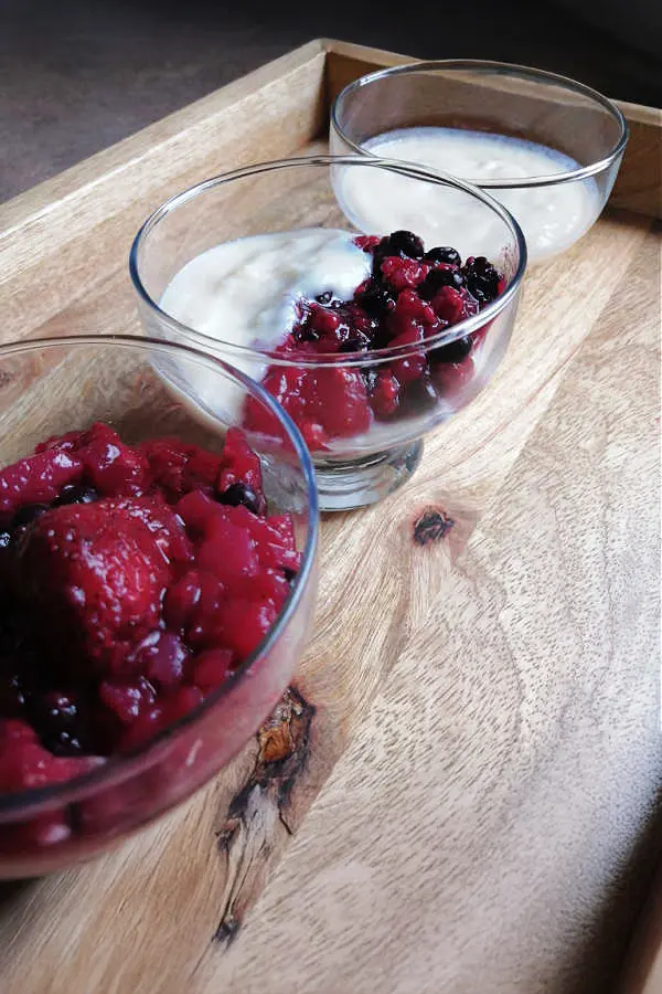 Three glass bowls on a wooden tray, each containing a berry dessert. The first and second bowls have mixed berries with vanilla cream, and the third contains vanilla cream.