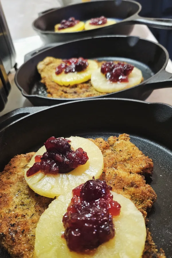 Breaded Hawaiian schnitzel topped with pineapple rings and whole-berry cranberry sauce in individual cast-iron pans, ready for the cheese