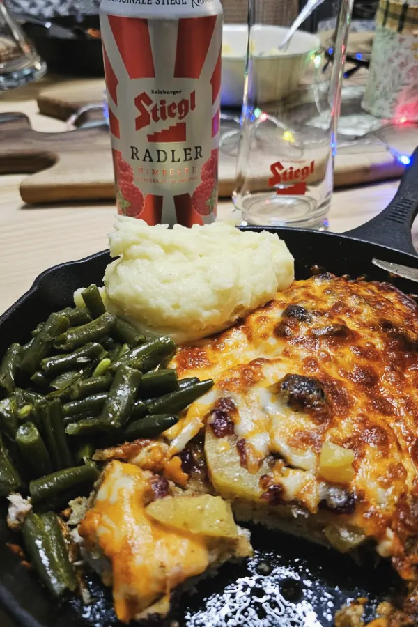 Hawaiian schnitzel with melted cheese and pineapple in a cast-iron pan, served with mashed potatoes, green beans and a raspberry Radler