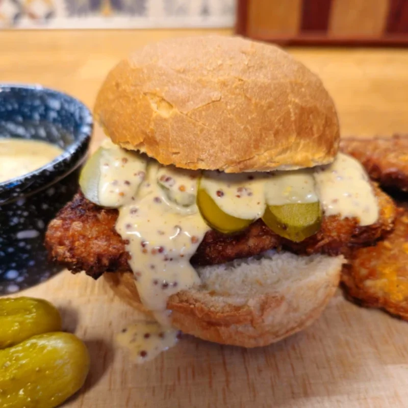 Pork schnitzel slider on a crusty roll topped with creamy beer mustard sauce and sliced pickles, served with extra sauce and mini pickles.
