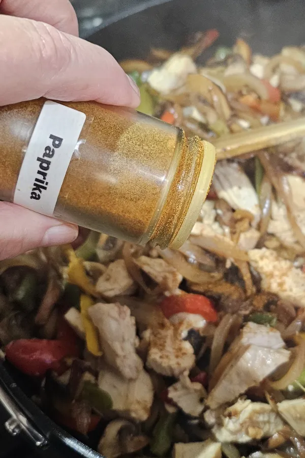 Hand sprinkling paprika from a jar over chicken, peppers and onions in a skillet