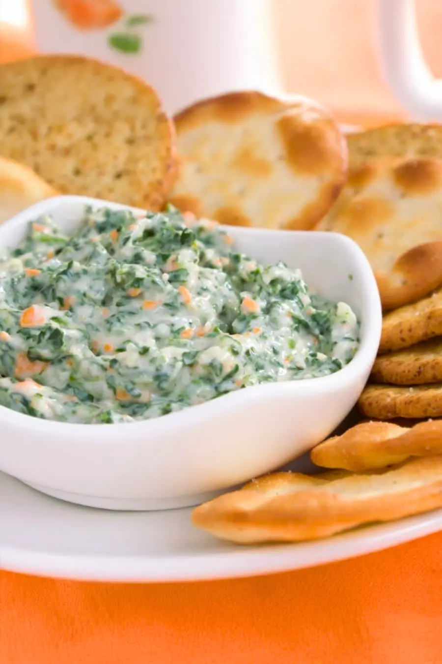 A white bowl filled with creamy spinach dip is surrounded by assorted crackers on a white plate.