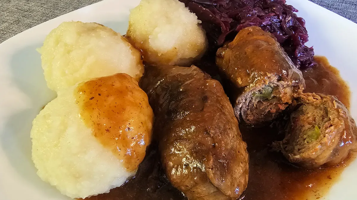 Beef rouladen with gravy, potato dumplings, and red cabbage on a white plate.