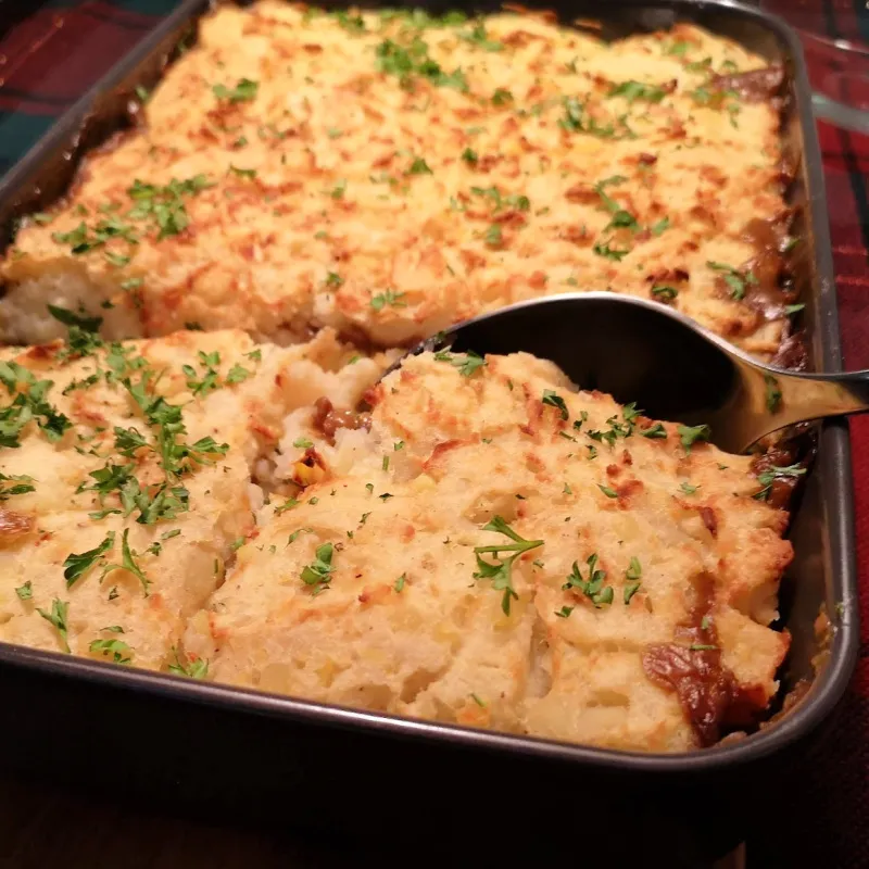 Vegan shepherd’s pie in a baking dish with a browned potato topping, parsley, and a serving spoon.