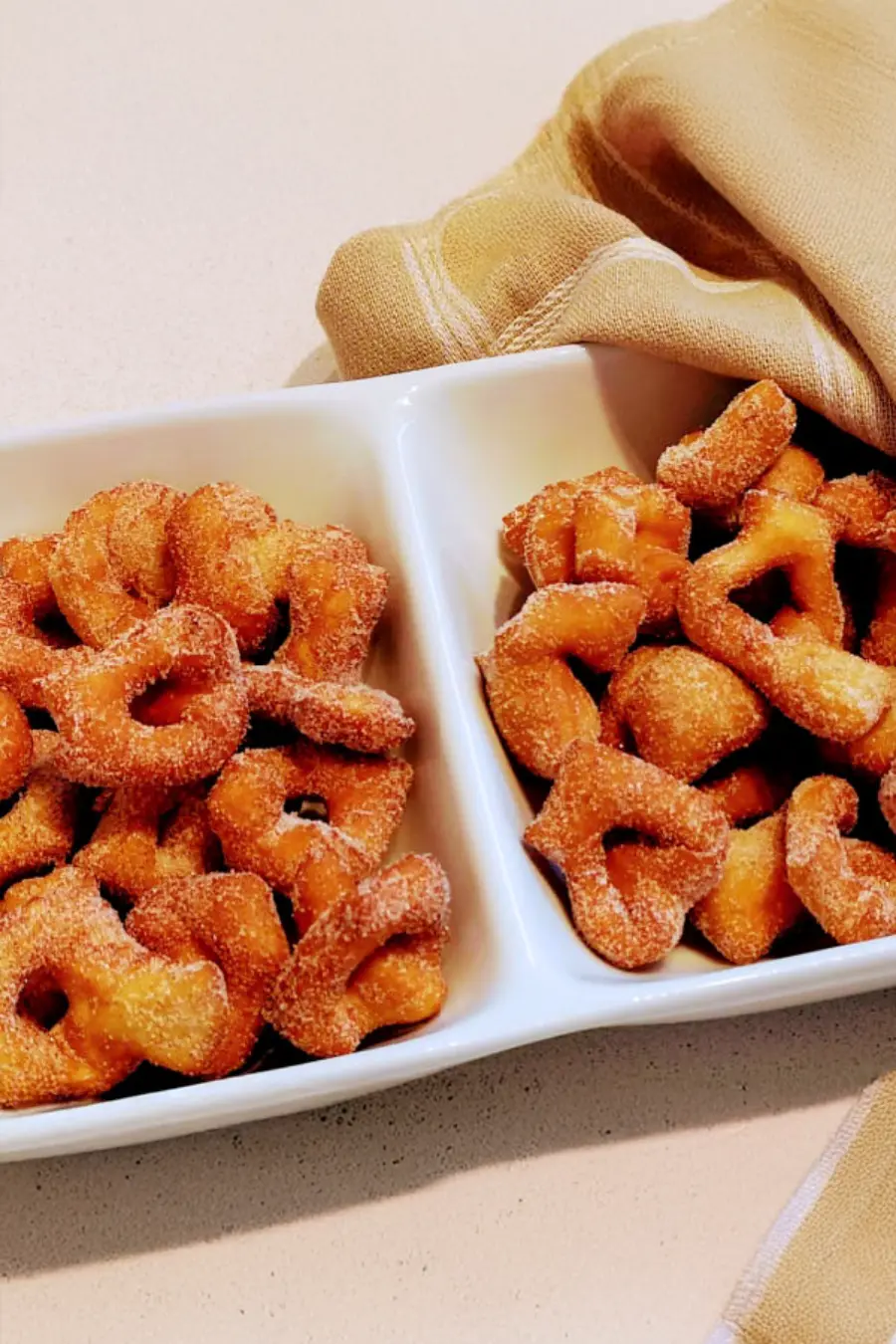 Two white dishes filled with sugared, fried donut-shaped pastries, placed on a light countertop with a tan cloth beside them.