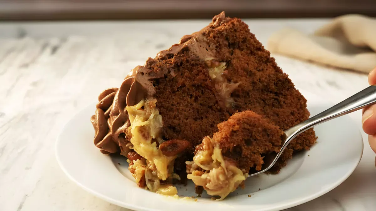 Slice of German chocolate cake on a white plate with coconut-pecan filling and chocolate frosting, with a forkful taken from the front