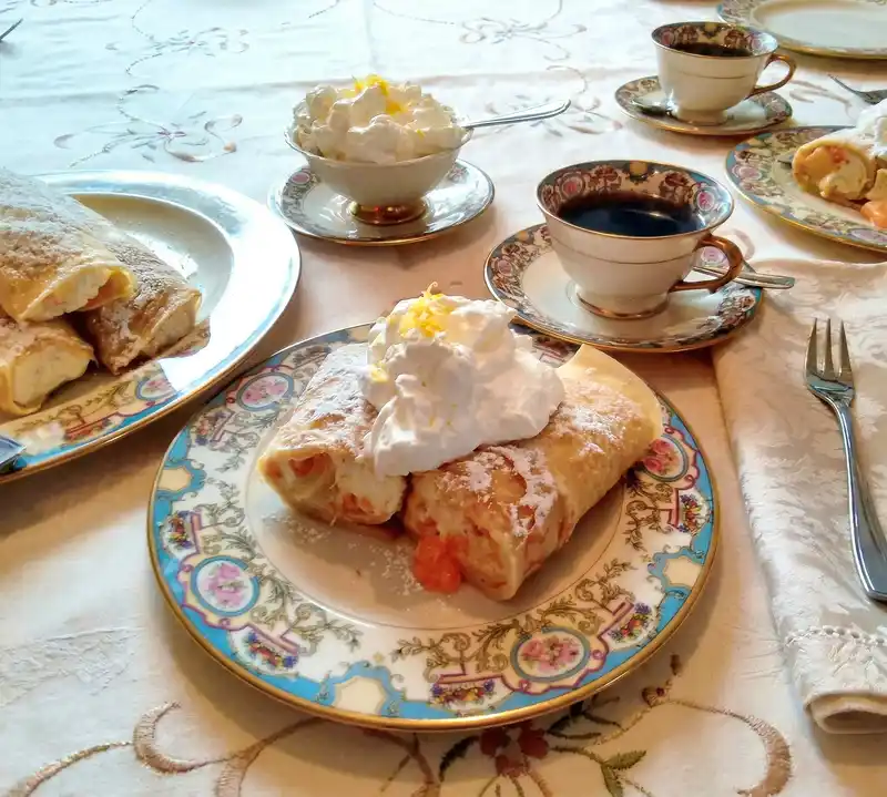 Delicate German-style crêpes filled with cream and fruit, dusted with powdered sugar and topped with whipped cream, served on fine china with a cup of black coffee.
