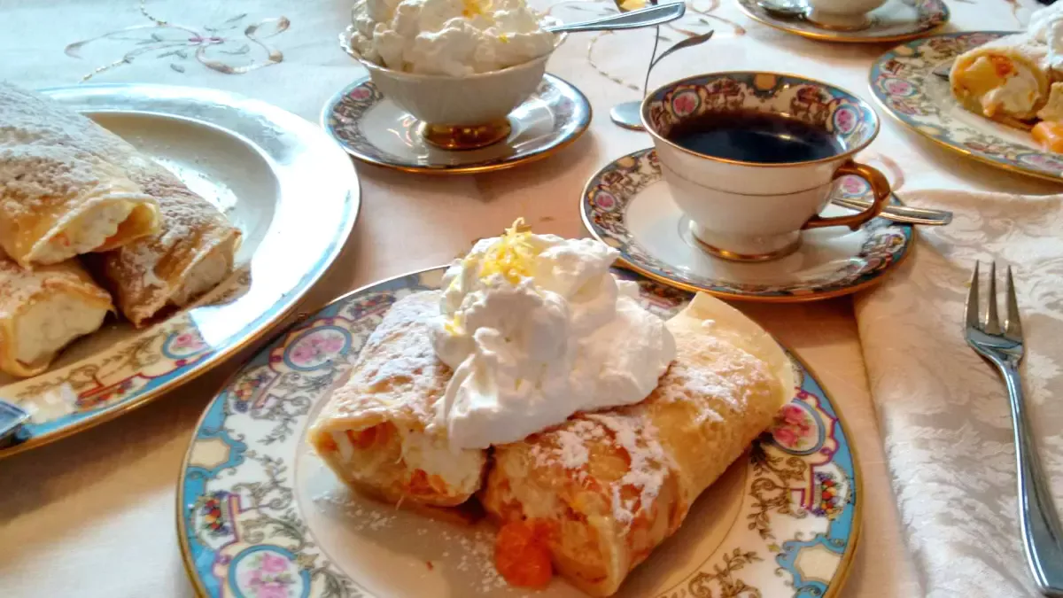 Delicate German-style crêpes filled with cream and fruit, dusted with powdered sugar and topped with whipped cream, served on fine china with a cup of black coffee.