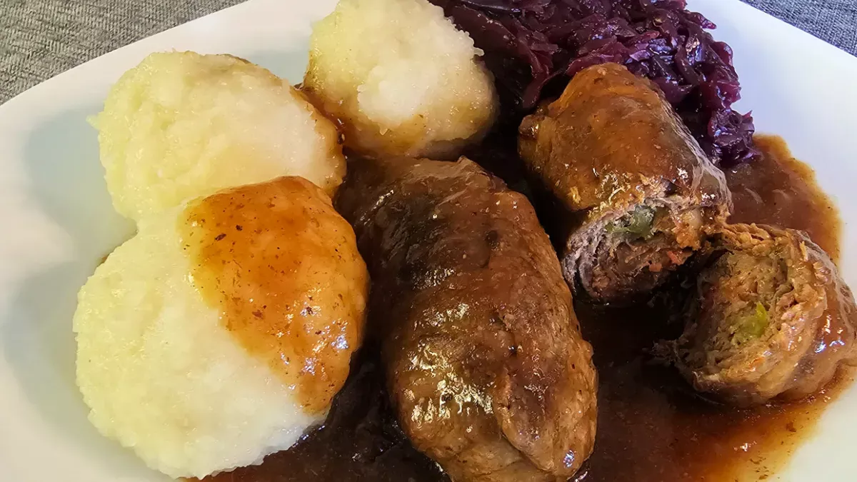 Beef rouladen with gravy, potato dumplings, and red cabbage on a white plate.
