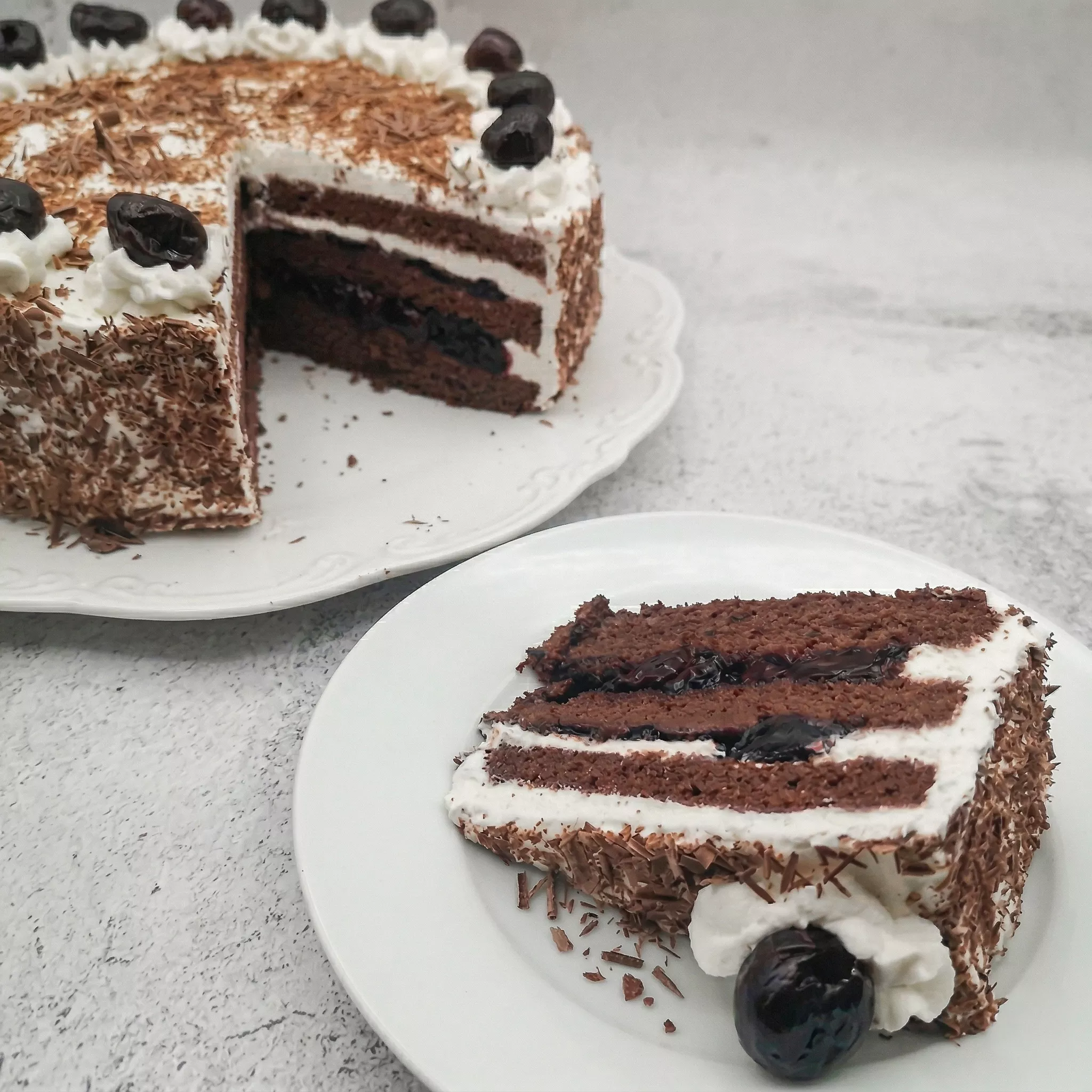 slice of traditional black forest cake on white plate with whole black forest cake in background